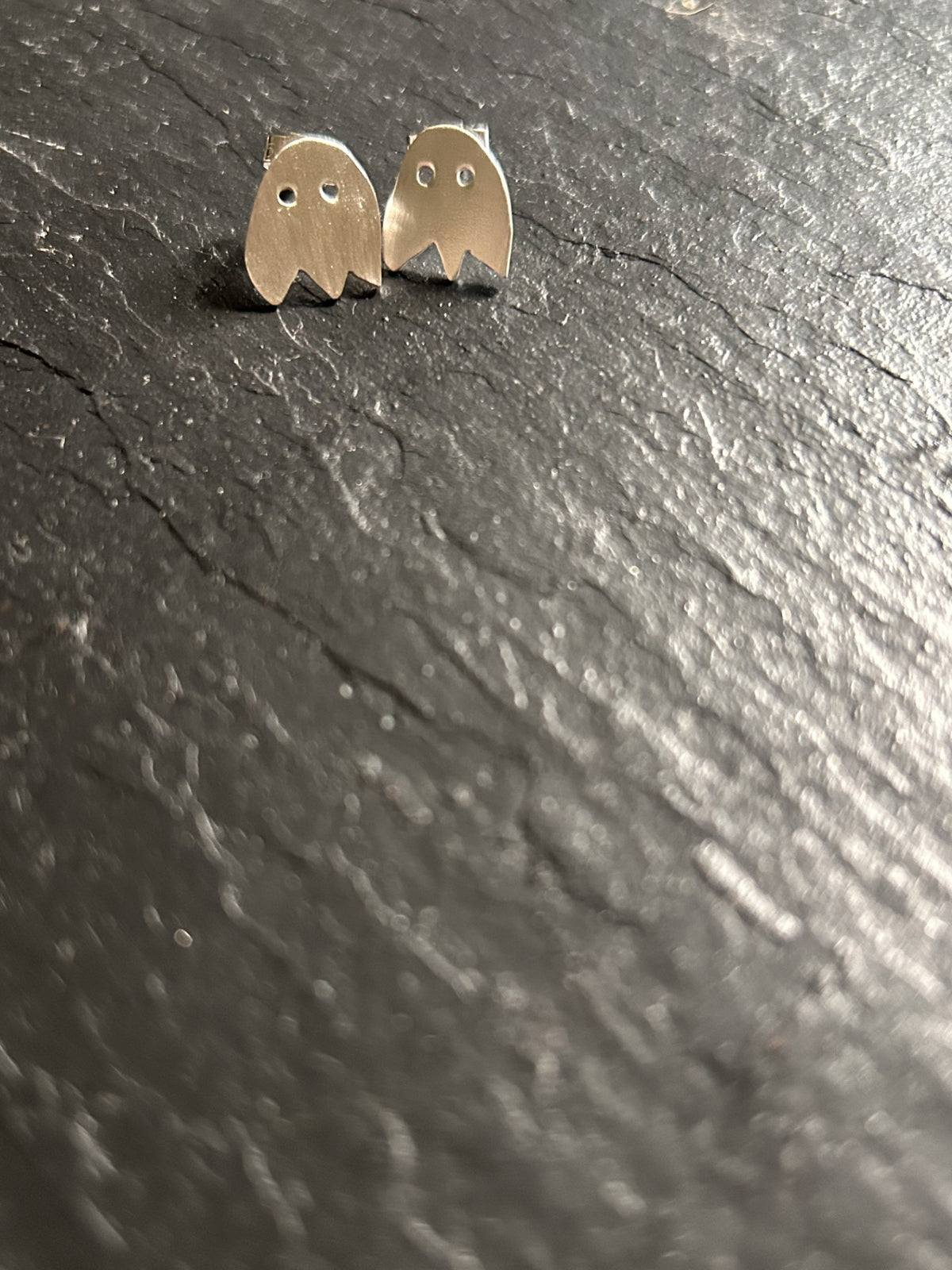 A pair of small ghost-shaped stud earrings in a silver-toned metal, displayed on a dark textured slate surface.